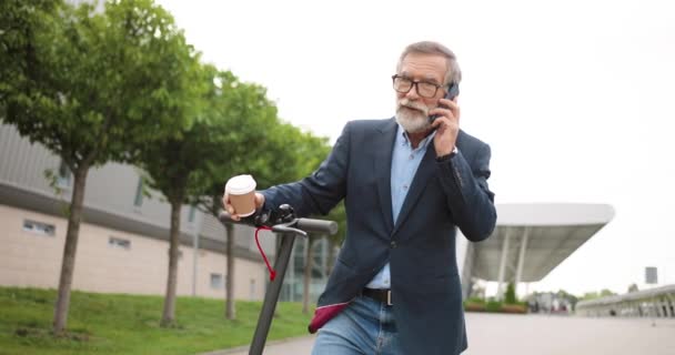 戴眼镜的老人站在镇上的电动车旁，举着咖啡，一边用手机交谈。老爷爷在户外用手机说话。打电话到街上。2.对话. — 图库视频影像