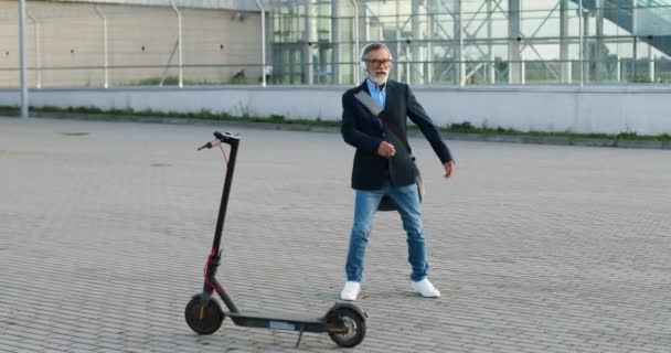 Hombre mayor en gafas y auriculares con bolsa de pie en scooter eléctrico en la calle urbana, escuchando música y bailando alegre y divertido. Viejo macho en airpods danza y moviéndose divertido fuera. — Vídeo de stock