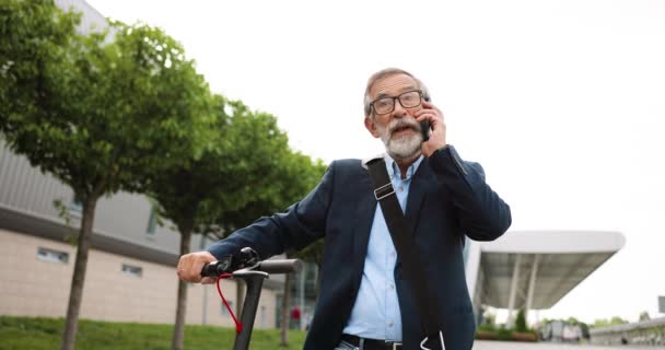 Ein älterer Mann mit Brille steht am Elektroroller in der Stadt und telefoniert. Alter Großvater telefoniert. Telefonat auf der Straße. Gespräch. — Stockvideo