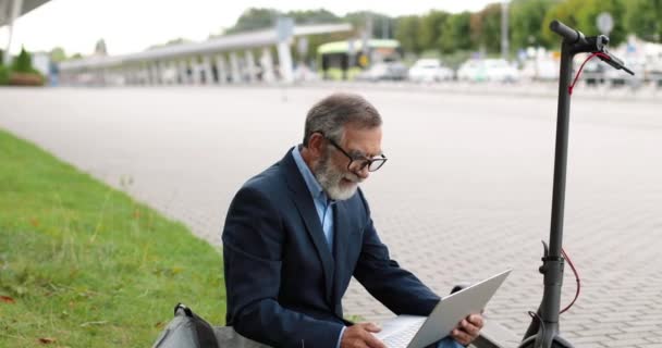 Gammal vit man i glasögon sitter utomhus på grönt gräs på elektrisk skoter och videochattar på bärbar dator. Man pratar via webbkamera på gatan. Farfar har videochatt och viftar med handen. — Stockvideo