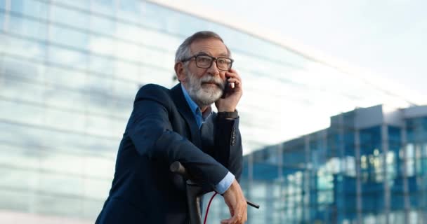 Homme âgé en lunettes debout à scooter électrique en ville et parlant au téléphone. Vieux grand-père parlant sur téléphone portable sur fond de bâtiment en verre moderne. Appel téléphonique à la rue. Conversation — Video