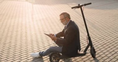 Gri saçlı, gözlüklü yaşlı adam caddede elektrikli scooter 'ın üzerinde oturuyor ve aletin bilgisayarına dokunuyor ya da kaydırıyor. Büyükbaba tablet aygıtı kullanıyor, internette geziniyor ve izliyor..