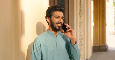 Hindu neşeli genç adam güneşli bir günde dışarıda duruyor ve cep telefonuyla konuşuyor. Hindistan 'dan yakışıklı bir adam cep telefonuyla konuşuyor ve gün ışığında gülümsüyor. Telefon görüşmesi kavramı.