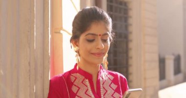 Alnında kırmızı nokta olan güzel, neşeli bir Hindu kadın. Geleneksel kıyafetiyle akıllı telefonları dinliyor ve mesaj yolluyor. Kadın cep telefonunda kayıyor. Açık havada. Mesajlaşma.