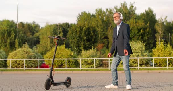 Senior man in bril en koptelefoon met tas staan op elektrische scooter op straat, luisteren naar muziek en dansen vrolijk en grappig. Oude grootvader in airpods dansen en bewegen grappig buiten. — Stockvideo
