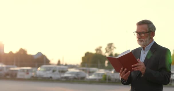 Viejo hombre de pelo gris caucásico en gafas caminando y leyendo libro al aire libre al atardecer en el aparcamiento de coches en el fondo. Profesor varón mayor paseando y leyendo un libro de texto. Estudiar. — Vídeo de stock