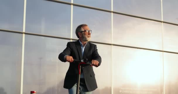 Portrait shot of Caucasian old man in glasses standing on electric scooter and looking at camera. Senior male pensioner or businessman on urban vehicle outdoors. Glass modern building on background. — Stock Video