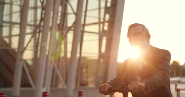 Retrato de anciano caucásico en gafas apoyadas en bicicleta y mirando a la cámara. Abuelo mayor en scooter eléctrico al aire libre. Hombre pensionista en gafas en bicicleta en la ciudad a la luz del sol al atardecer. — Vídeo de stock