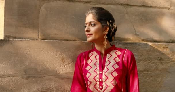 Портрет індуїстської молодої красивої жінки з крапкою на лобі і червоним традиційним одягом, що обертається обличчям і посміхається камері. На вулиці. Досить чарівна щаслива дівчина з Індії біля антикварної стіни на вулиці. — стокове відео