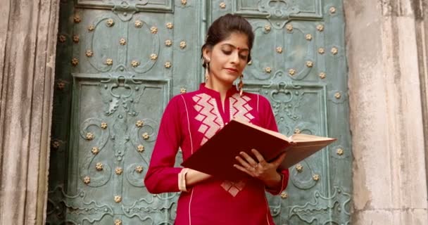 ドアの外に立ってカメラに笑みを浮かべて額にドットと美しい若いヒンドゥ教徒の女性の肖像画。読書本。美しいですスタイリッシュな魅力的な女性で赤伝統的な衣装とともに教科書. — ストック動画