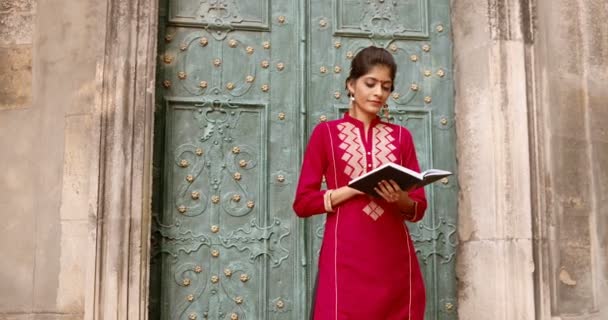 Портрет красивої молодої індуської жінки з крапкою на лобі, що стоїть на вулиці і посміхається до камери. Читаю книжку. Досить стильна чарівна жінка в червоному традиційному одязі з підручником. — стокове відео