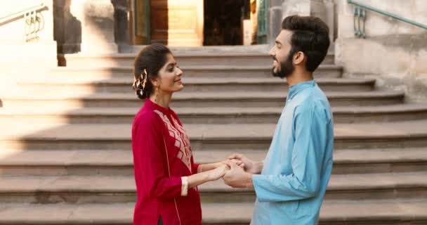 Hindu beautiful young woman and handsome man in traditional outfits standing outdoor at steps of building, holding hands and looking at each other with smile and love. Romantic relationships. — Stock Video