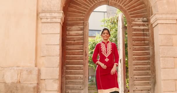 Retrato de hindu bela jovem em roupa tradicional em pé ao ar livre na rua em porta de madeira e posando para câmera. Modelo feminino alegre atraente na Índia. Menina bonita em vestido vermelho. — Vídeo de Stock