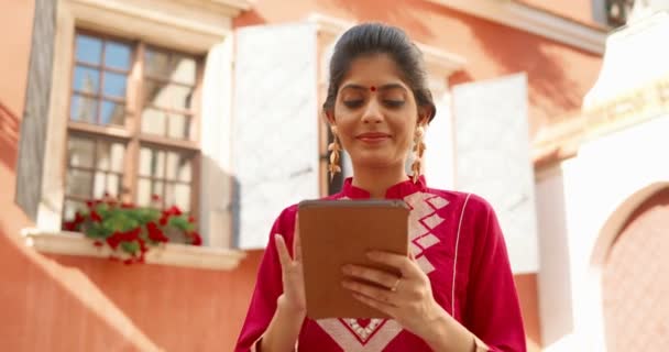 Hindú hermosa sonrió mujer de pie al aire libre en la calle en el día soleado y ver algo en el dispositivo de la tableta. Chica de la India usando la computadora gadget, tocando y desplazándose. Navegación femenina online. — Vídeo de stock