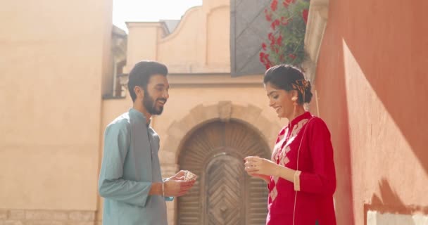 Mujer joven hindú y el hombre en trajes tradicionales de pie al aire libre en el hermoso patio, sonriendo, hablando y bebiendo de copas. Hombre y mujer bebiendo té o café en una cita romántica y charlando. — Vídeos de Stock