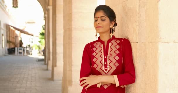 Portret hinduskiej młodej pięknej kobiety z kropką na czole i w czerwonym tradycyjnym stroju, opartej na ścianie i uśmiechniętej do kamery. Dość uroczy uśmiechnięta radosna dziewczyna z Indii na zewnątrz. — Wideo stockowe