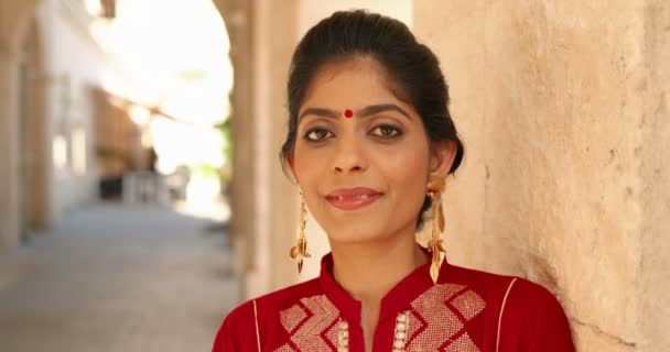 Згорніть індуїстську молоду вродливу жінку з крапкою на лобі й червоним традиційним одягом, спираючись на стіну й усміхаючись до фотоапарату. Портрет чарівної усмішки радісна дівчина з Індії.. — стокове відео