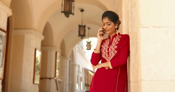 Mujer hermosa joven hindú con punto en la frente y en traje tradicional rojo, apoyada en la pared y hablando por teléfono celular. Chica bastante sonriente hablando en el teléfono móvil al aire libre. Llamada telefónica. — Vídeos de Stock