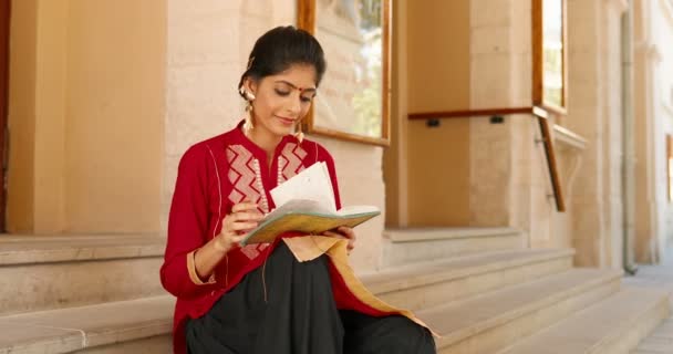 Wanita Hindu muda yang cantik dengan pakaian tradisional merah dan dengan titik di dahi duduk di tangga luar ruangan dan membaca catatan di buku catatan. Perempuan dari India memeriksa perencana. Perencanaan hari. — Stok Video