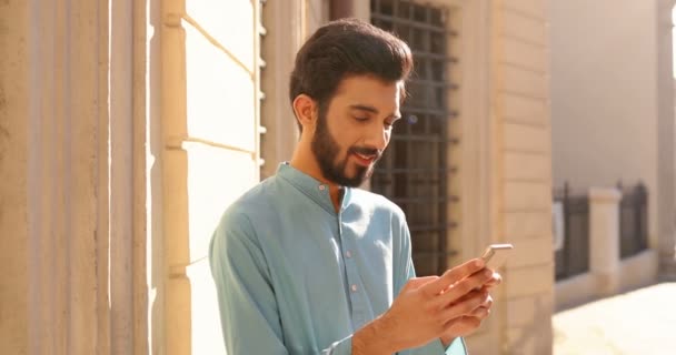 Geleneksel kıyafetli Hindu bir gencin portresi caddede duruyor, akıllı telefondan mesaj atıyor ve kameraya gülümsüyor. SMS gönderiliyor. Telefonunu dinleyip kaydıran bir adam. Açık havada erkek mesajları.. — Stok video
