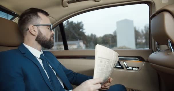 Kaukázusi fiatal, jóképű üzletember szemüvegben, öltönyben és nyakkendőben ül egy drága autó hátsó ülésén, és munkába megy reggel. Férfi híreket olvas. Férfi utas olvasni újságot az autóban. — Stock videók