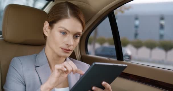 Close up de jovem bela mulher caucasiana sentado no banco de trás do carro caro e mensagens de texto no dispositivo tablet. Mulher de negócios muito bem sucedida tocando e rolando no computador gadget, indo para o trabalho. — Vídeo de Stock