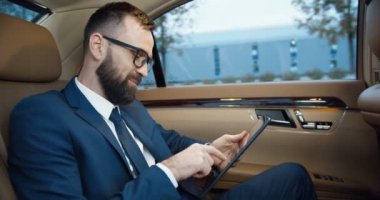 Gözlüklü yakışıklı beyaz adam pahalı arabanın arka koltuğunda oturuyor ve tablet cihazıyla mesaj yazıyor. Başarılı, neşeli iş adamı, iş yerinden başlayarak alet bilgisayarını tıklatıyor ve kaydırıyor..