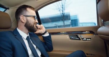 Gözlüklü yakışıklı beyaz adam pahalı bir arabanın arka koltuğunda oturuyor ve cep telefonuyla konuşuyor. Başarılı neşeli iş adamı mobil telefondan konuşuyor. Telefonunuz var. İş görüşmesi.