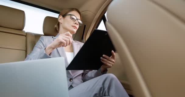 Gözlüklü beyaz bir iş kadını arabanın arka koltuğunda oturuyor ve belgelerle uğraşıyor. İş hayatı yaşayan bir kadın işe giderken belgelerini ve sözleşmelerini kontrol ediyor. Kadın avukat evrakları hazırlıyor.. — Stok video