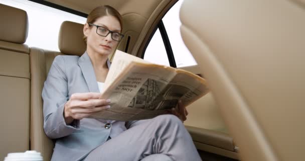 Caucasian beautiful businesswoman in glasses sitting on backseat of expensive car and going to work in the morning. Woman sipping hot drink and reading news. Female drinking coffe and read newspaper. — Stock Video