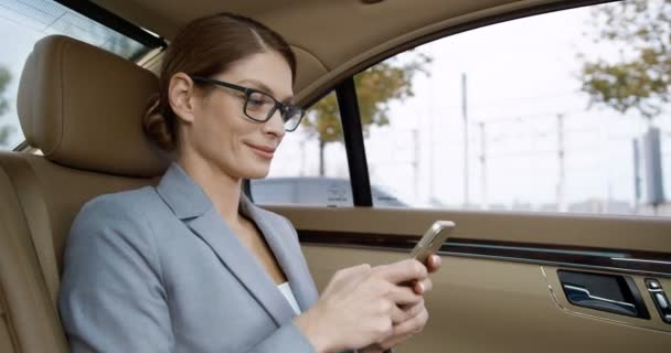 행복 한 젊고 아름다운 백인 여성이 값비싼 차 뒷자리에 앉아 스마트폰으로 웃으며 문자 메시지를 보내고 있습니다. 꽤 사업적 인 여성이 핸드폰으로 창을 두드리고 검색하는 모습. — 비디오