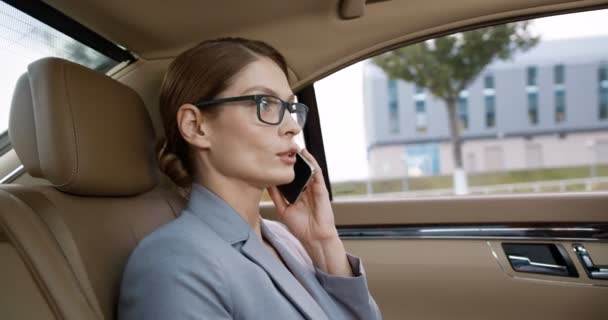 Pahalı bir arabanın arka koltuğunda oturan ve cep telefonuyla konuşan gözlüklü güzel beyaz kadını yakından çek. Başarılı bir iş kadını cep telefonuyla konuşuyor. Telefonunuz var. İş görüşmesi — Stok video