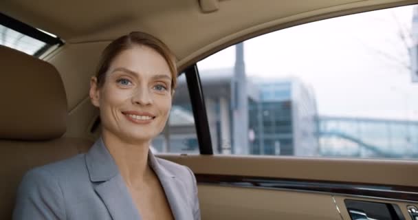 Retrato de la joven mujer de negocios hermosa caucásica girando cara a cámara y sonriendo sinceramente mientras se sienta en un coche caro en el asiento trasero. Mujer en estilo de negocios con sonrisa va a trabajar por la mañana — Vídeo de stock