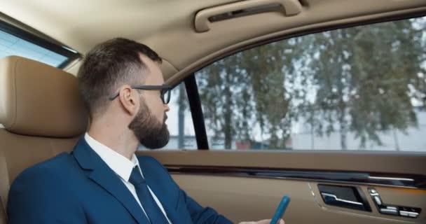 Takım elbiseli, kravat ve gözlüklü yakışıklı beyaz adam pahalı bir arabanın arka koltuğunda oturuyor ve elinde cep telefonu var. Sakallı ve cep telefonlu ciddi bir işadamı, bir yere gidiyor ve pencereye bakıyor.. — Stok video