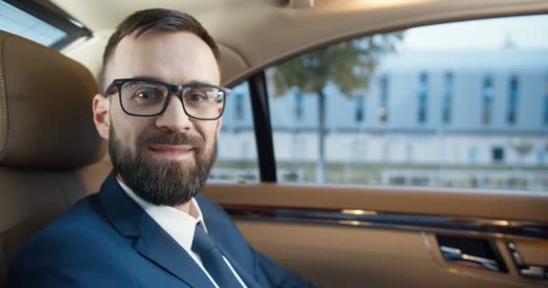 Ritratto di bell'uomo d'affari caucasico in occhiali, vestito e cravatta guardando la macchina fotografica e sorridendo sinceramente, seduto in auto costosa sul sedile posteriore. Uomo in stile business andare a lavorare in ingorgo. — Video Stock