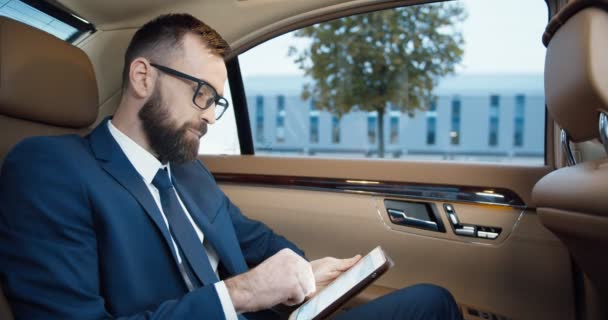高価な車の後部座席に座って眼鏡でハンサムな白人男性とタブレットデバイス上のテキストメッセージ。スーツやネクタイで成功したビジネスマンは、仕事から行く、ガジェットのコンピュータ上でタップしてスクロールします. — ストック動画