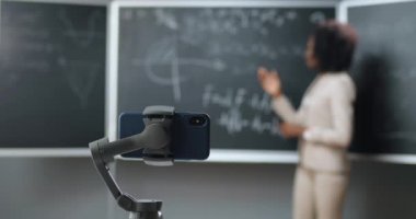 Cep telefonunun okuldaki webcam 'deki çevrimiçi dersinin video kaydı. Kara tahtada matematik ya da fizik formülleri öğreten gözlüklü Afrikalı Amerikalı kadın öğretim görevlisi. Tecrit konsepti. Salgın Çalışması.