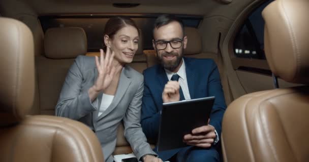 Kafkasyalı işadamı ve iş kadını arabanın arka koltuğunda oturup tablet cihazlı web kamerasıyla görüntülü sohbet ediyorlar. Birkaç iş ortağı gülümsüyor ve görüntülü sohbet ediyor. Görüntülü arama. Çevrimiçi sohbet — Stok video