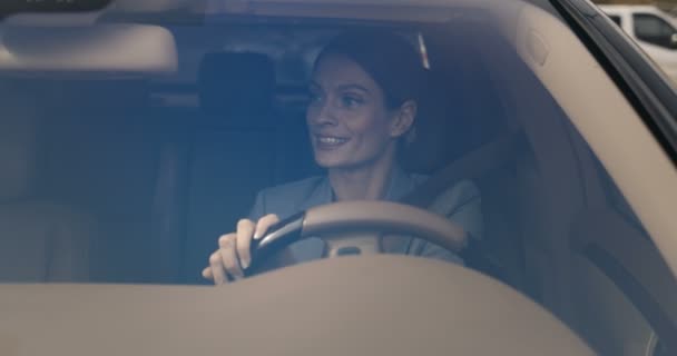 Vista através do pára-brisas. Bonita sorriu caucasiana jovem empresária sentado no volante, usando cinto de segurança e condução de carro caro. Atraente alegre motorista feminino transformando. — Vídeo de Stock