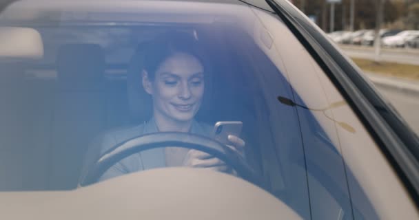 Hermosa joven caucásica feliz mujer sentada al volante y mensaje de texto en el teléfono inteligente. Bonita sonrisa conductor femenino tocando y desplazándose en el teléfono móvil. Vista a través del parabrisas. — Vídeos de Stock