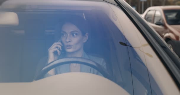 Linda jovem caucasiana feliz mulher sentada no volante e falando no celular. Bonito sorriso motorista feminino falando no telefone celular. Vista através do pára-brisas. Conversa telefónica. — Vídeo de Stock