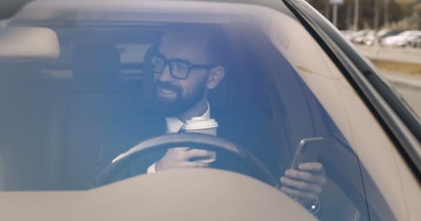 Όμορφος επιχειρηματίας κάθεται στο τιμόνι και γραπτών μηνυμάτων σε smartphone, ενώ πίνοντας ποτό. Άντρας οδηγός σε γυαλιά πατώντας και κύλιση στο κινητό τηλέφωνο, πίνοντας καφέ. Μέσω ανεμοθώρακα. — Αρχείο Βίντεο