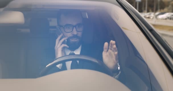 Bel giovane uomo d'affari caucasico seduto al volante e che parla al cellulare. Autista di sesso maschile che parla al cellulare e guarda l'orologio. Vista attraverso il parabrezza. Essere in ritardo. Conversazione. — Video Stock