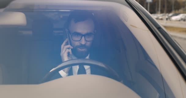 Biały, wściekły biznesmen siedzący za kierownicą i rozmawiający przez komórkę. Mężczyzna mówi przez telefon komórkowy i kończy rozmowę w gniewie. Widok przez przednią szybę. Problemy. — Wideo stockowe