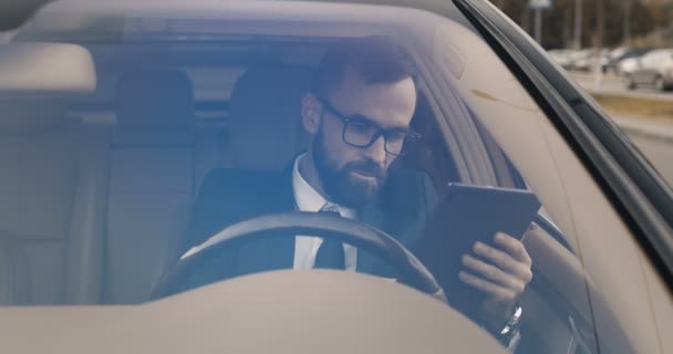 Bel homme caucasien assis au volant et textos sur tablette dispositif. Souriant mâle conducteur tapant et défilant en ligne sur l'ordinateur gadget. Par le pare-brise. Recherche dans la carte pour l'itinéraire. — Video