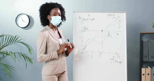 Joven mujer afroamericana profesora de máscara médica de pie a bordo en el aula y diciendo leyes de física o geometría a la clase. Concepto pandémico. Escuela durante coronavirus. Lección educativa. — Vídeos de Stock