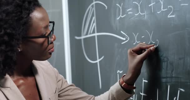 Kara tahtaya formüller ve mahematik yasalar yazan Afrikalı Amerikalı genç bayan öğretmeni kapatın. Okul konsepti. Fizik kanunlarını açıklayan gözlüklü kadın öğretim görevlisi.. — Stok video