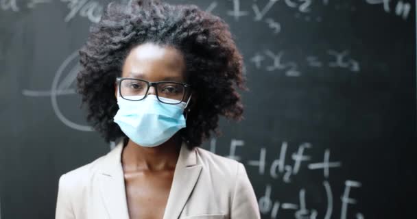 Porträt einer jungen afroamerikanischen Lehrerin mit Brille und medizinischer Maske, die im Klassenzimmer in die Kamera blickt. Tafel mit Formeln im Hintergrund. Coronavirus-Konzept. Pandemische Schulbildung. — Stockvideo