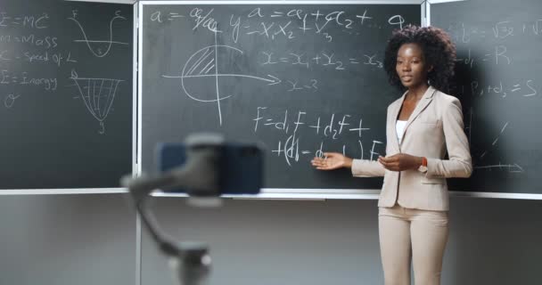 Grabación de vídeo de Smartphone lección virtual en webcam en la escuela. Estudiando en línea. Profesora afroamericana que enseña matemáticas o fórmulas físicas en pizarra. Concepto de bloqueo. Estudio pandémico. — Vídeo de stock