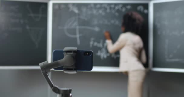 Mobiltelefon felvétel online video lecke az iskolában. A lezárás elszigetelt tanulás. Afrikai amerikai tanárnő elmagyarázza a matematikai vagy fizika képleteket a táblán. Járványügyi oktatás. — Stock videók